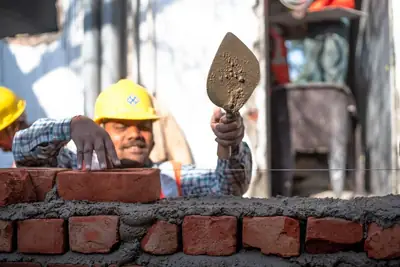 A mason in a yellow helmet laying a brick on a mortar bed