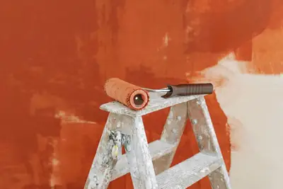 A paint roller resting on a stepladder against a half-painted orange wall